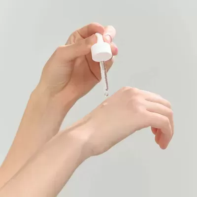 Mãos a aplicar líquido transparente com conta-gotas branco na pele