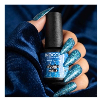 Frasco de verniz INOCOS azul glitter segurado por mão com unhas compridas azuis glitter em fundo azul