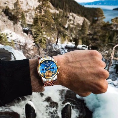 Relógio ORBUS azul com bracelete metálica em pulso, fundo de floresta e neve