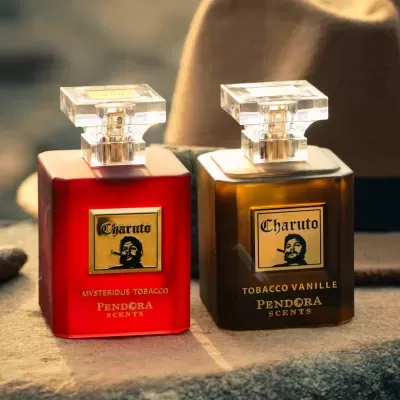 Duas garrafas de perfume Charuto com etiquetas douradas, uma vermelha e outra castanha, sobre uma pedra com um chapéu castanho ao fundo