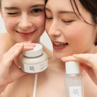 Duas mulheres segurando produtos cosméticos em frascos com rótulos em inglês e coreano
