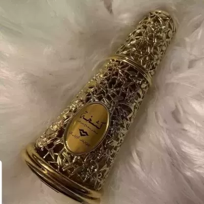 Frasco decorativo dourado com padrão floral e texto em árabe sobre fundo branco felpudo