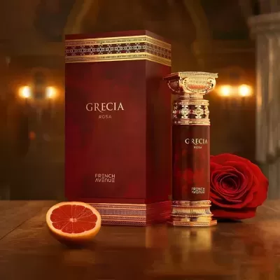 Frasco de perfume e caixa vermelhos com detalhes dourados e texto GRECIA ROSA e FRENCH AVENUE, rosa vermelha e grapefruit lado a lado na mesa
