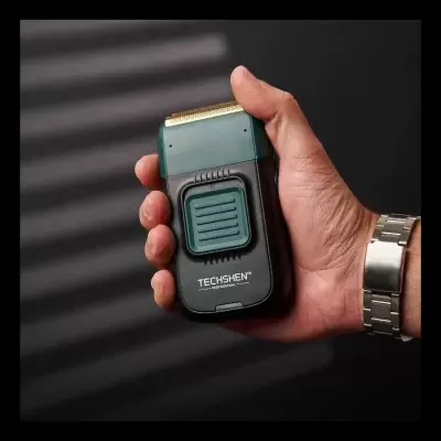 Barbeador elétrico preto e verde com detalhes dourados na lâmina e texto TECHSHEN PROFESSIONAL segurado por uma mão com pulseira metálica