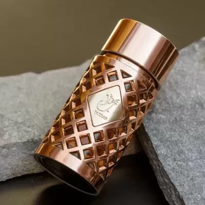 Frasco de perfume metálico cobre com padrão geométrico e etiqueta com texto árabe