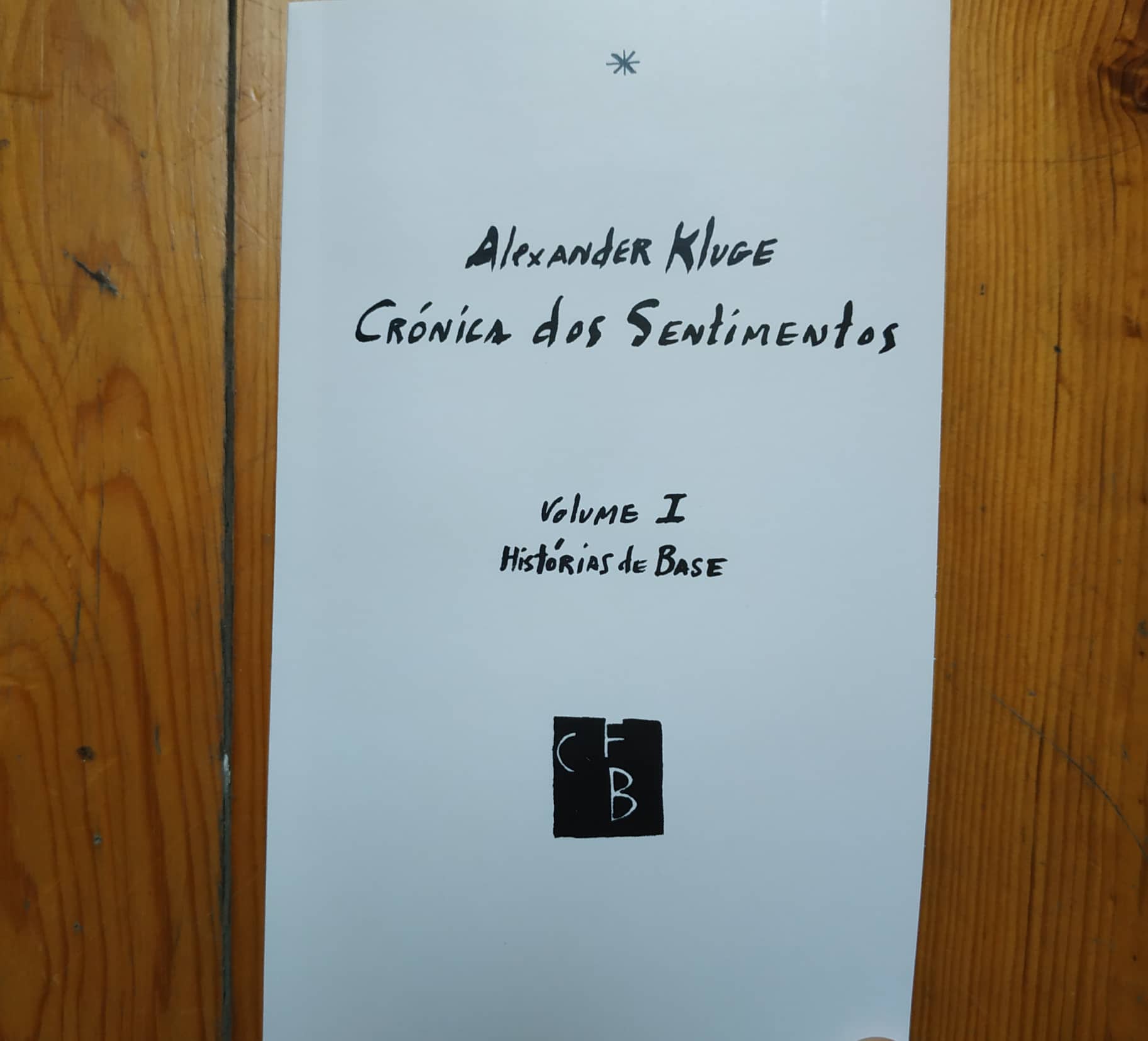 Crónica dos Sentimentos Vol.I - Histórias de Base Capa branca de livro com título escrito à mão e logotipo CFB sobre madeira.
