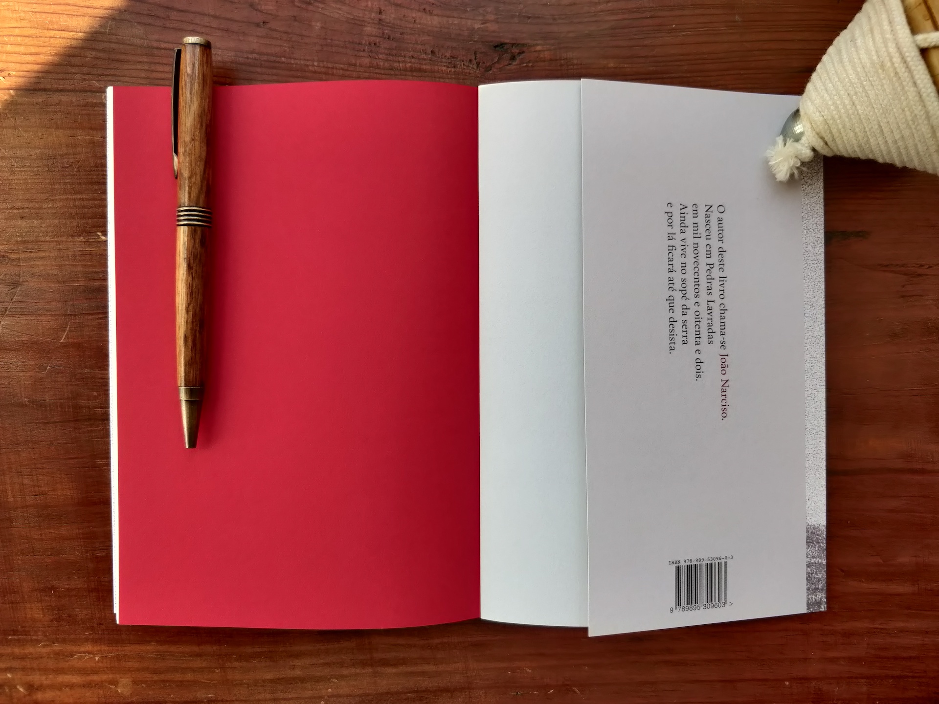 estes ventos negros Livro aberto com uma caneta de madeira sobre uma página vermelha, texto e código de barras na página oposta, sobre mesa de madeira e cesto têxtil ao lado.