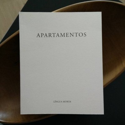 Livro branco com título APARTAMENTOS e texto LÍNGUA MORTA sobre superfície escura e objeto de madeira.