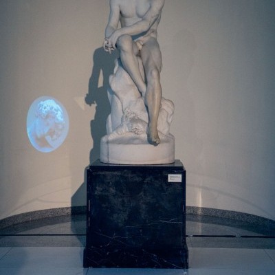 Estátua de mármore branco de figura humana sentada sobre pedestal negro