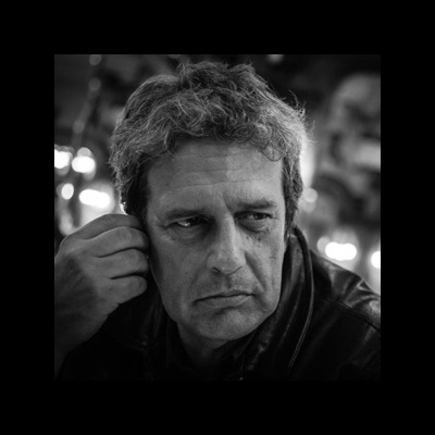 Homem de meia-idade com casaco de pele escuro e expressão séria em fundo desfocado