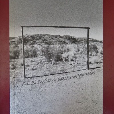 Fotografia preto e branco de uma estrutura retangular de madeira em terreno natural com texto no chão