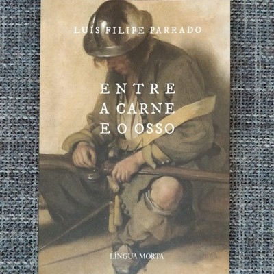 Capa de livro com homem com chapéu e roupa antiga sentado, título 'ENTRE A CARNE E O OSSO' e autor Luiz Filipe Parrado