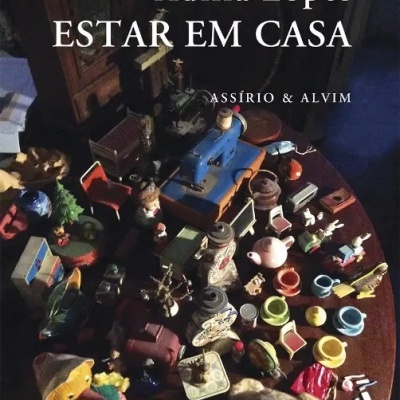 Capa de livro com o título 'Adília Lopes ESTAR EM CASA' e objetos variados em miniatura sobre uma mesa.