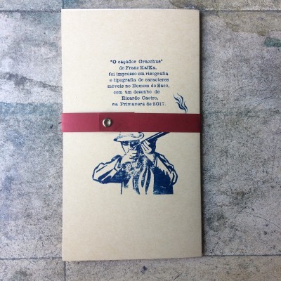 Folheto com capa bege, ilustração azul de homem com arma, texto azul e faixa vermelha com ilhó