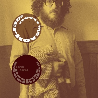 Homem com camisa bege e óculos segurando manta de malha em ambiente com parede amarela