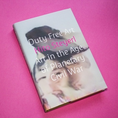 Livro com capa com texto Duty Free Art Hito Steyerl sobre fundo desfocado, colocado numa superfície rosa.