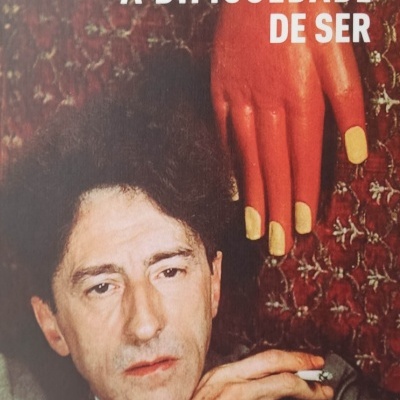 Capa do livro 'A Dificuldade de Ser' de Jean Cocteau com homem a segurar cigarro e mão gigante laranja ao fundo