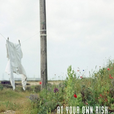 Roupa branca pendurada em estendal ao lado de poste de madeira num campo com vegetação e flores, texto VINTAGE JARMAN e AT YOUR OWN RISK.