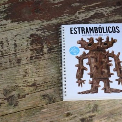 Livro Estrambólicos com figura abstrata castanha sobre mesa de madeira envelhecida