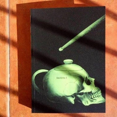 Livro com capa preta e ilustração verde de crânio em forma de bule sobre chão de azulejos alaranjados iluminado pelo sol.