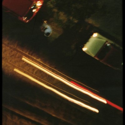 Estrada noturna com dois carros estacionados, luzes de veículos e pessoa na calçada