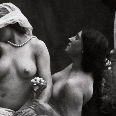 Duas mulheres semi-nuas em pose clássica a preto e branco, uma com véu e colar de pérolas, segurando flores.