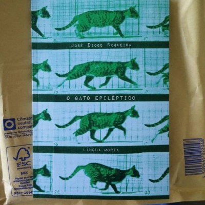 Livro com capa azul mostrando imagens de gato verde em várias posicoes e texto com titulo e autor em branco sobre envelope castanho.