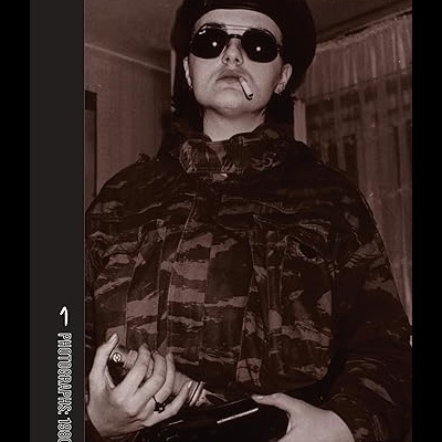 Pessoa com casaco militar, boina preta e óculos escuros segurando objeto escuro