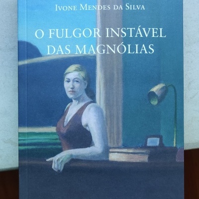 Capa de livro azul com pintura de mulher loira ao lado de mesa e candeeiro, texto O Fulgor Instável das Magnólias.