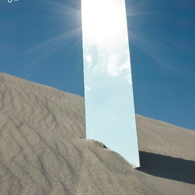 Espelho retangular refletindo o sol numa duna de areia sob céu azul claro com texto branco