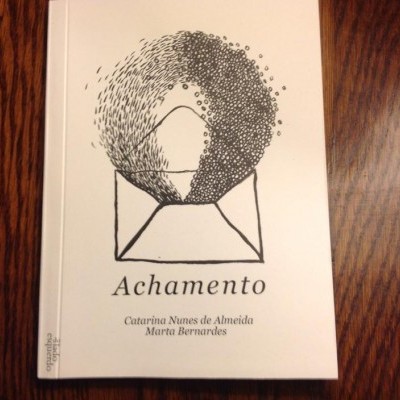 Livro branco com ilustração abstrata preta e texto 'Achamento' em cima de uma mesa de madeira