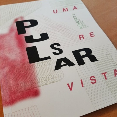 Capa de publicação branca com texto em preto e vermelho com as palavras PULSAR, UMA, RE, VISTA, NUMÉRO 2, sobre mesa de madeira.