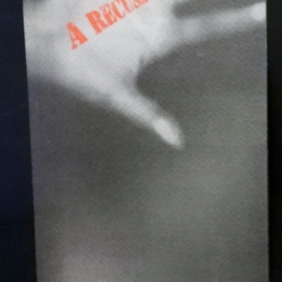 Capa de livro cinzenta com mão desfocada e texto A RECUSA em vermelho