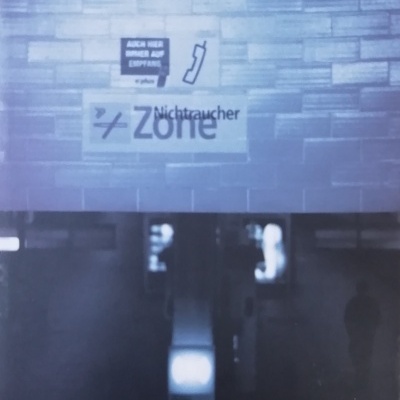 Corredor com painéis texturizados, letreiros com texto bilingue e sinais em alemão