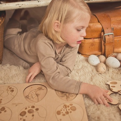 Criança loira a brincar com puzzle de madeira sobre tapete felpudo beige junto a ovos decorativos e mala castanha