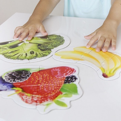 Puzzles infantis de alimentos com brócolos, bananas e frutos vermelhos sobre mesa branca.