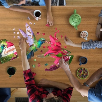 Pessoas a montar puzzle de beija-flor e flores numa mesa com caixa do puzzle, canecas e bule verde.