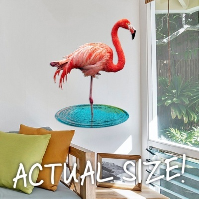 Adesivo de parede de flamingo em posição de equilíbrio com texto 'ACTUAL SIZE!' numa sala com sofá e almofadas.