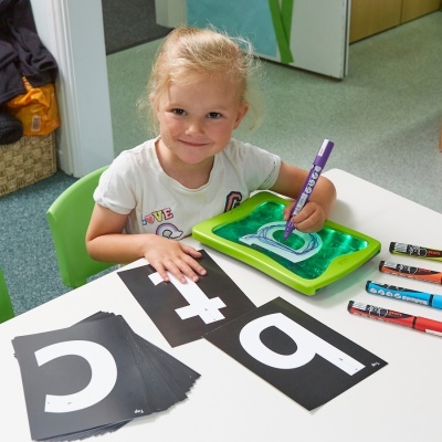 Menina pintando letra numa bandeja verde com cartões pretos e brancos numa mesa branca