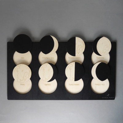 Jogo de memória de peças redondas em madeira clara com texto e ilustrações, peças pretas a cobrir algumas, numa base preta sobre fundo cinza.