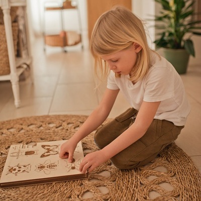 Criança sentada em tapete trançado beige a manusear livro de madeira com desenhos de insetos