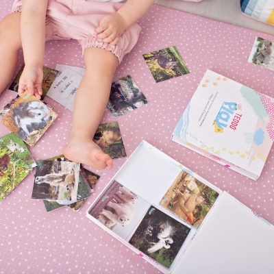 Criança sentada em tecido cor-de-rosa com cartas fotográficas de animais espalhadas e caixa aberta