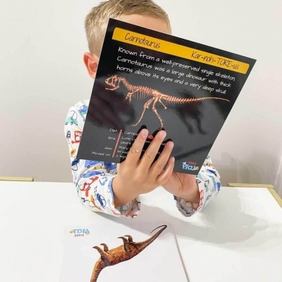 Criança com pijama colorido segurando livro sobre dinossauro Carnotaurus com esqueleto e texto em inglês