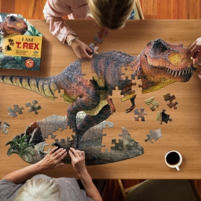 Quebra-cabeças de dinossauro T-Rex sendo montado por duas pessoas numa mesa de madeira com café e caixa do jogo