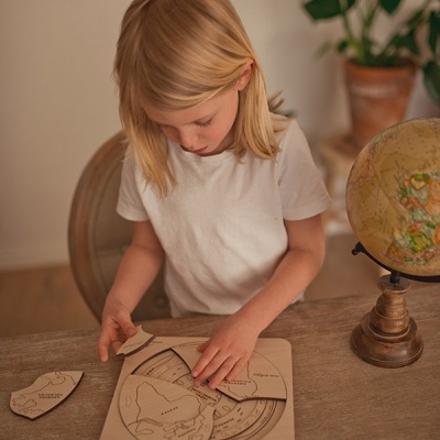 Criança a montar puzzle de madeira com mapas numa mesa de madeira, com globo e planta ao fundo