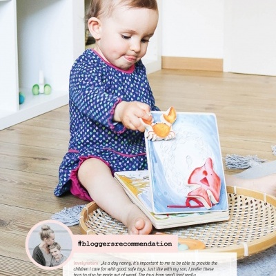 Bebé sentado no chão de madeira a brincar com livro ilustrado e brinquedo de raposa, perto de estante branca e cesta de palha