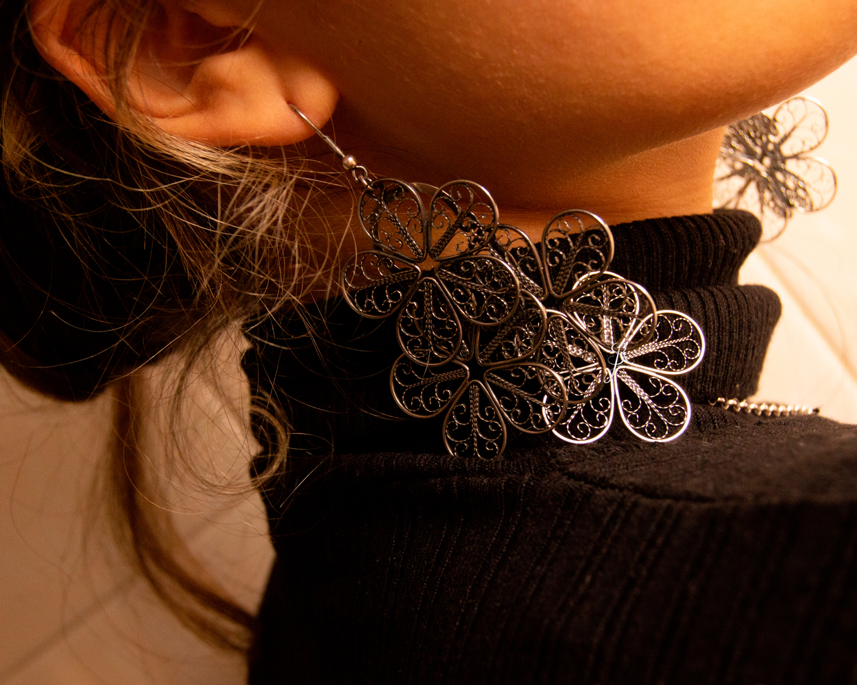 Brincos Flores de Filigrana Brincos longos prateados em forma de flores, usados por pessoa com suéter preto de gola alta