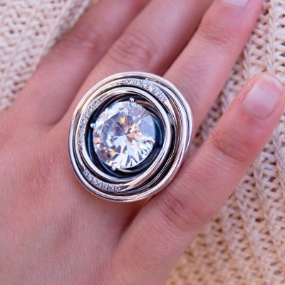 Anel de metal prateado com grande pedra redonda e transparente num dedo sobre tecido bege de malha