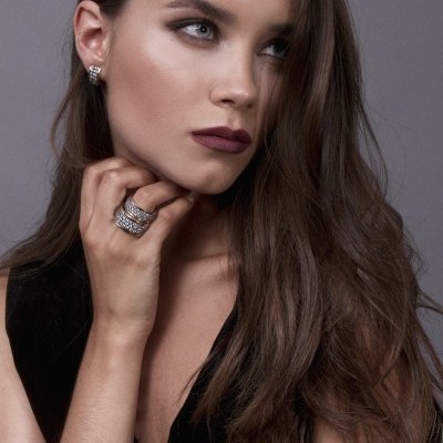 Mulher com brincos e anéis prateados com pedras brilhantes, roupa preta e fundo cinzento