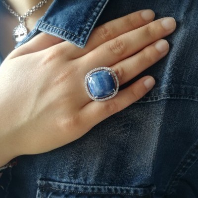 Anel com pedra azul e cristais usado em mão sobre camisa de ganga azul.